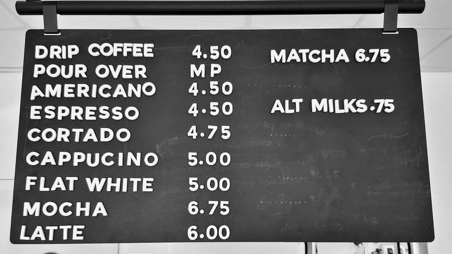 A black and white photo of a hanging menu board at a coffee shop with white block letters on a black eraser board.