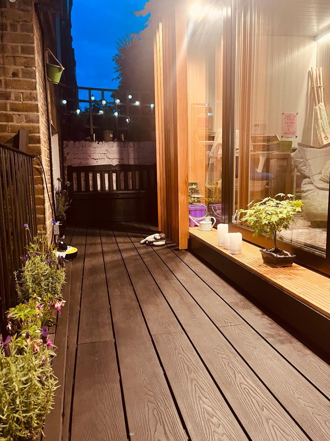 A warm evening view of a modern garden pod with glass doors glowing from within. The pod opens onto a wooden deck lined with plants and fairy lights, creating a serene and inviting writing space.