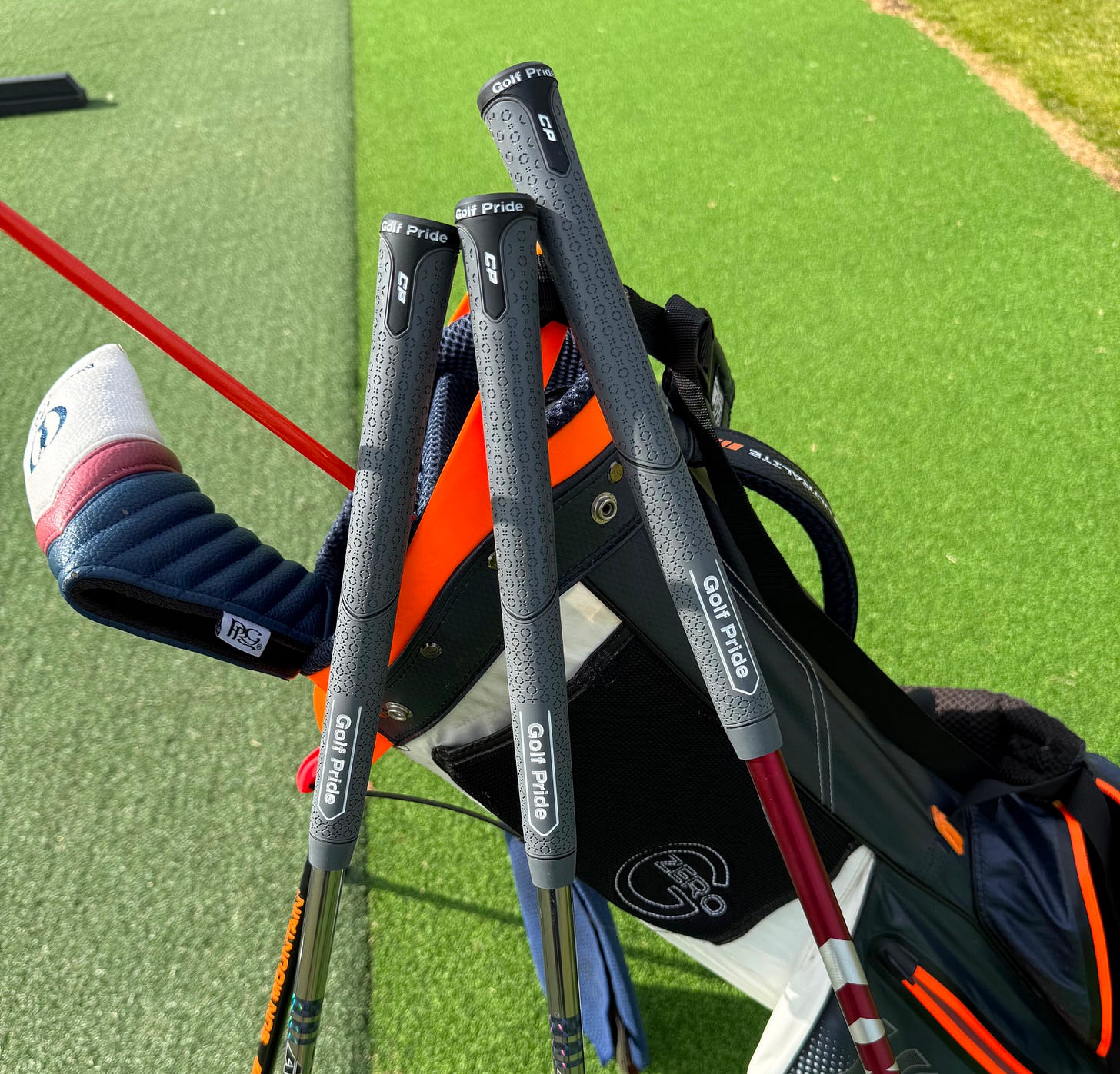 Three grey Golf Pride CP midsize grips fitted to golf clubs beside a golf bag on a practice ground. Three grey Golf Pride CP midsize grips fitted to golf clubs beside a golf bag on a practice ground.