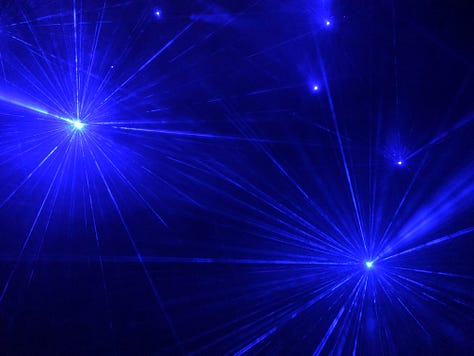 A blue and violet laser light beams through the dark forest. All around, sapphire faerie lights shine starbursts into the night