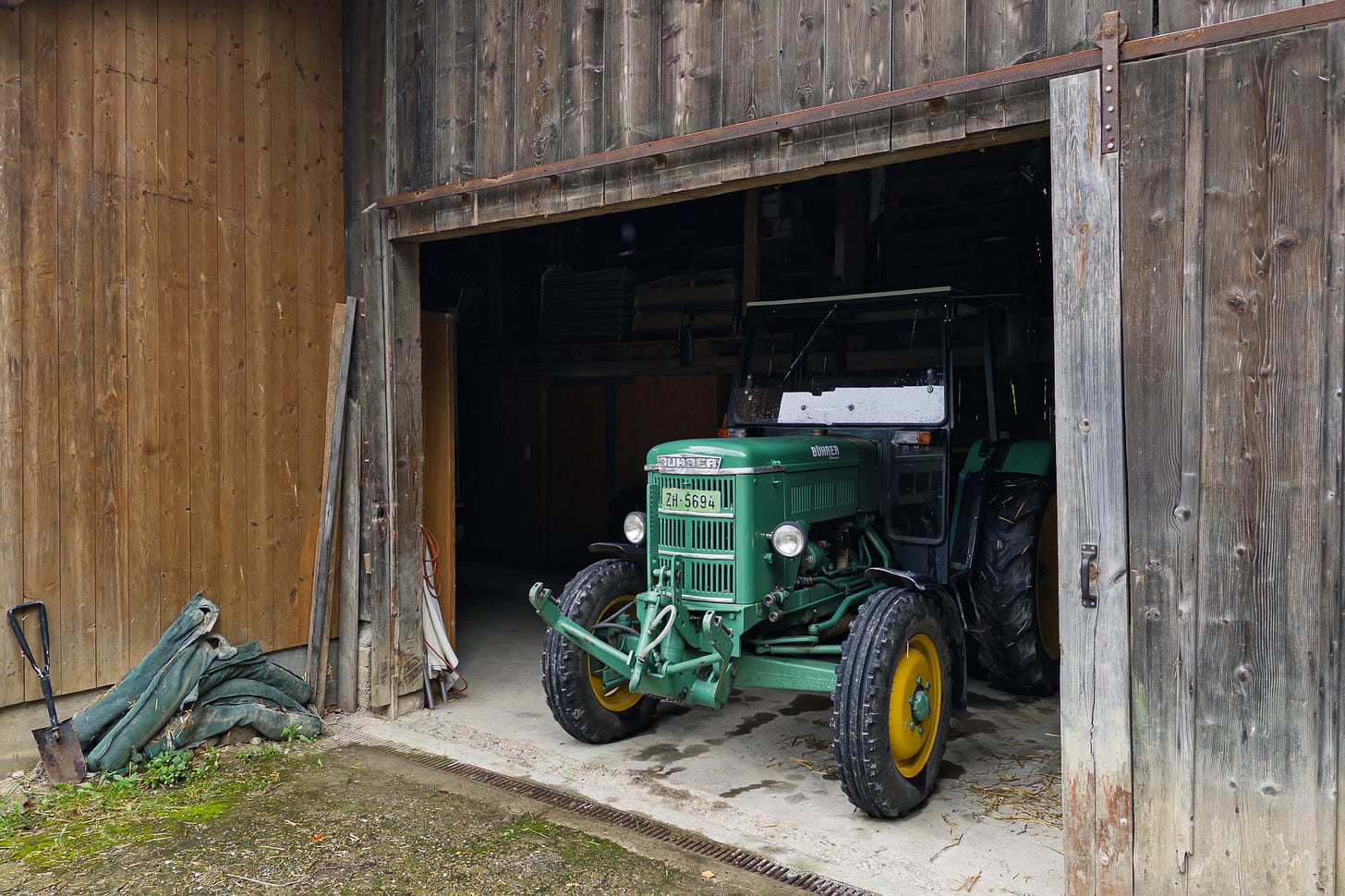 Ein grüner Bührer-Traktor mit Baujahr 1964 steht halb in einer geöffneten Holzscheune. Der kleine Oldtimer mit gelben Felgen und Zürcher Nummernschild ist frontal leicht schräg zu sehen. Ein grüner Bührer-Traktor mit Baujahr 1964 steht halb in einer geöffneten Holzscheune. Der kleine Oldtimer mit gelben Felgen und Zürcher Nummernschild ist frontal leicht schräg zu sehen.