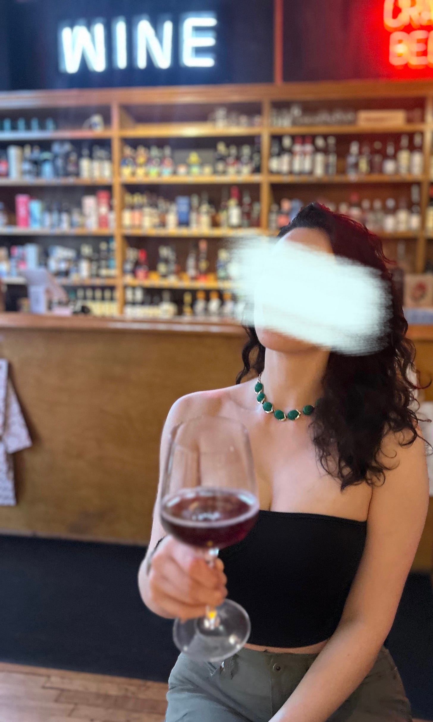 Shot of Elena at a wine bar. In the foreground, Elena is sitting and wearing a black tube top showing just a peek of clevage and belly, paired with army green pants and a green/gold choker necklace. Her long dark hair is down and wavy, and swepy to the side. The wine glass she's toasting with has a semi-transparent red wine, a sparkling rosé that was sweet but not in a cloying way. She was impressed! The background is the wine shop, and it's out of focus. There is a cash register counter, and behind that is a light-wood shelf with various liquor and wine bottles for sale. The shelf is packed, as it should be. Above that, you can see the dark blue walls with two neon signs. One is light blue and reads "Wine," and the other is red and reads "Draft Beer". Great spot.