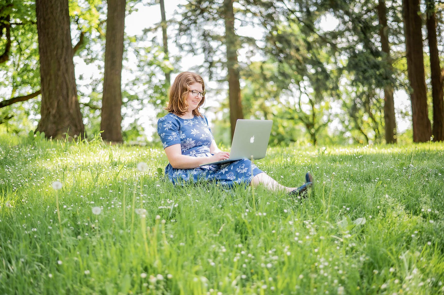 Me, a white woman with red hair sitting with a laptop across my legs. Sitting in the grass with dandelion puffs and surrounded by trees and warm spots of sunshine.