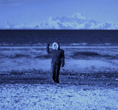 On a metallic blue background facing the rise of mountains, a lone figure in a parka, waves.