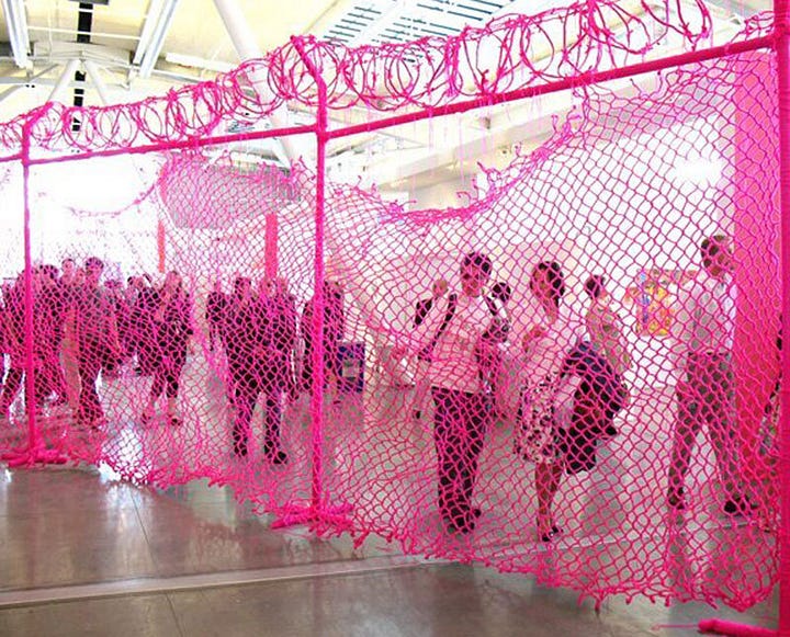 1) A large knit pink chain-link fence with knit barbed wire is seen with people outside of it, looking in. 2) The fence is now outside, in an alleyway, with a graffiti-ed wall to the left.
