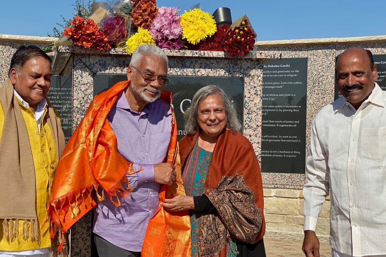 Dr Carson stands with Ela Gandhi in front of a large marble memorial to her grandfather, Mahatma Gandhi. They wear orange woven shawls. There are bouquets of flowers lying atop the marble stone behind them.