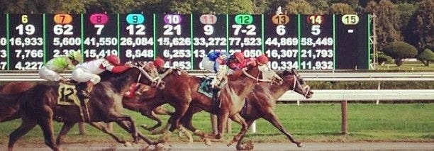 This may contain: jockeys racing their horses down the track at a horse race course in front of score boards