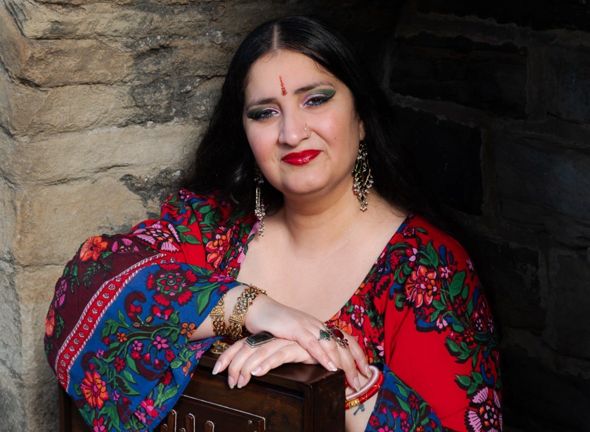 A photo of British-Asian musician Satnam Galsian. She is wearing traditional Punjabi dress, and her arms are resting on a shruti box.