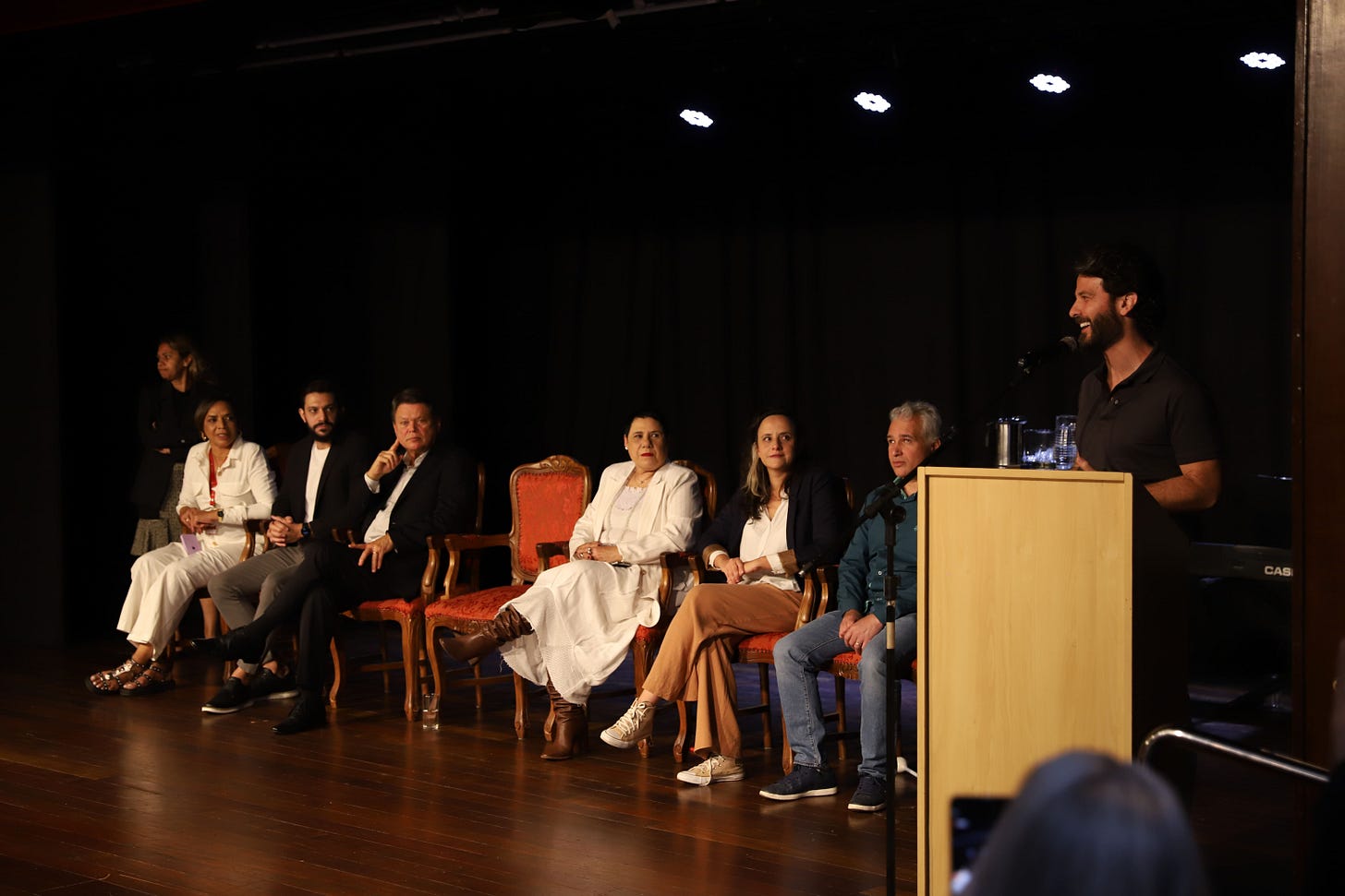 #descrevemospravocê  Em um auditório, sete pessoas participam de um evento no palco. Seis delas estão sentadas em cadeiras de madeira com estofado laranja, dispostas lado a lado, enquanto um homem, à direita da imagem, está de pé atrás de um púlpito de madeira clara, falando ao microfone e sorrindo. O grupo é composto por homens e mulheres com idades variadas, vestindo trajes sociais ou semiformais em cores neutras como preto, branco, azul e bege. Ao fundo, uma cortina preta cobre toda a parede, e acima há fileiras de luzes de palco acesas. O piso é de madeira escura polida. No canto esquerdo, uma mulher em pé observa a cena, afastada do grupo principal. No canto inferior direito, parcialmente desfocado, aparece o topo da cabeça de uma pessoa que fotografa ou filma o momento com um celular.