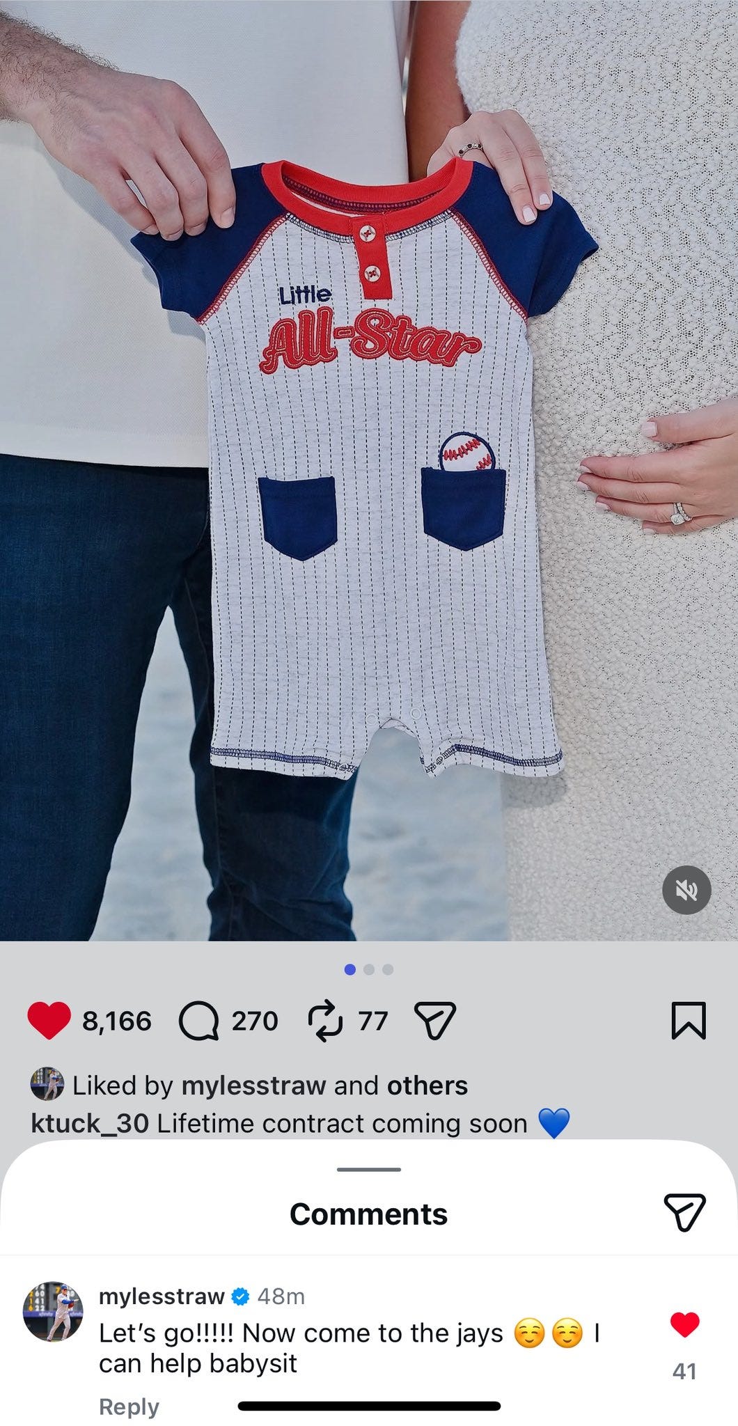 A man and a woman stand side by side holding up a small white baby onesie resembling a baseball uniform. The onesie has short sleeves in blue and red with Little All-Star text across the chest and a Toronto Blue Jays logo on the pocket. The onesie includes a small pocket and snap closures at the bottom. The background is plain white.