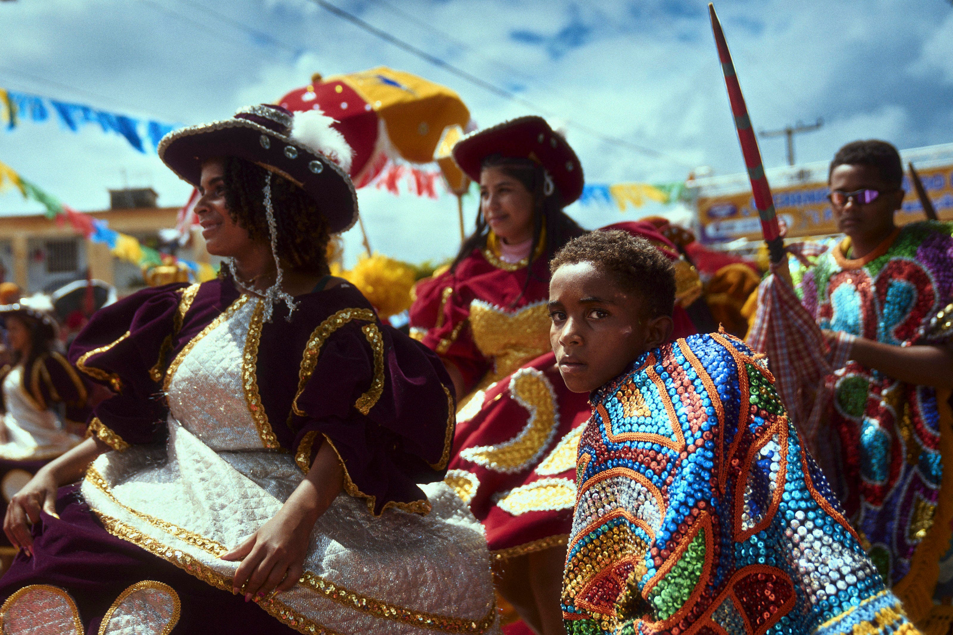 People in colorful, ornate costumes participate in a festive outdoor parade under blue skies, with vibrant banners and umbrellas decorating the background. A young boy in beaded attire looks toward the camera.