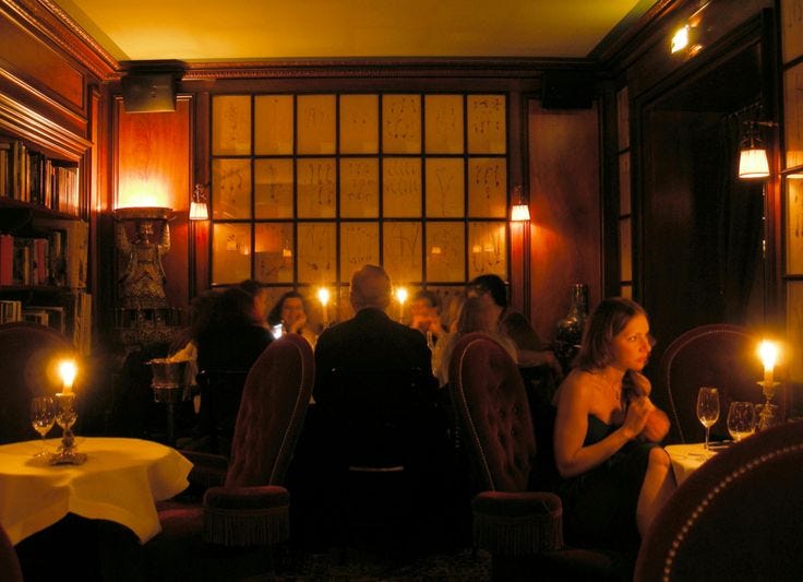 This may contain: people sitting at tables in a dimly lit restaurant with candles on the table and bookshelves behind them