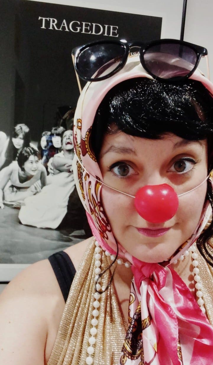 a white woman in a black wig with a clown nose, a pink headscarf and sunglasses stands in front of a black and white poster with the word 'tradegie'