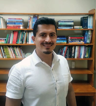 Um homem sorridente de cabelos escuros e cavanhaque posa para a foto usando uma camisa de manga curta branca. Ele está posicionado à frente de uma estante de madeira clara repleta de livros coloridos, organizados em fileiras. O ambiente tem uma iluminação suave e profissional, sugerindo um escritório ou biblioteca.