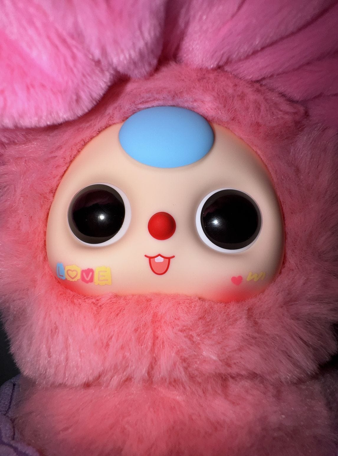 Extreme close-up of a pink Baby Three plush doll: big black eyes ringed in white, red button nose, tiny smiling mouth, blue circle on the forehead, and a cheek decal spelling “LOVE” in colorful blocks, all framed by soft faux-fur. 