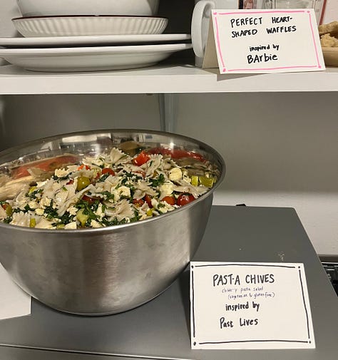 A bowl of pasta salad labeled "Pasta Chives", a Miller High Life for The Holdovers, two tall boys labeled "Anatomy of a Tall Boy," a sign reading "Jeffrey Bites - mac & cheese bites and caramelized onion & feta bites."