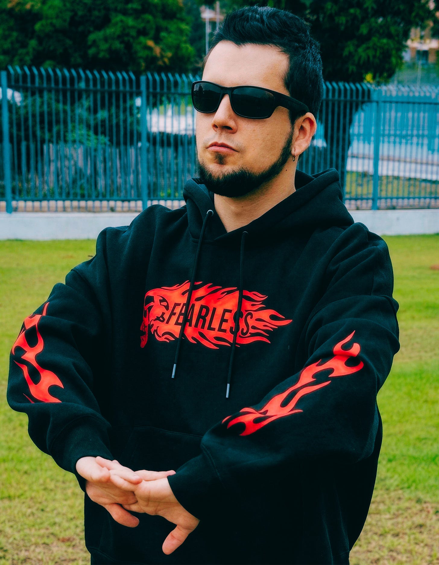 Pedro Neto stands outdoors in a park, arms crossed at the waist, wearing sunglasses and a black hoodie printed with a red flame design and the word FEARLESS. A teal metal fence and trees are behind him. Pedro Neto stands outdoors in a park, arms crossed at the waist, wearing sunglasses and a black hoodie printed with a red flame design and the word FEARLESS. A teal metal fence and trees are behind him.