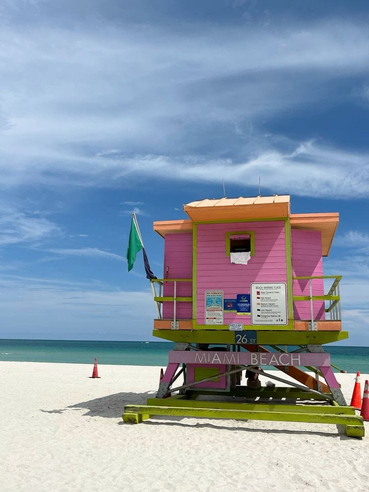 A vibrant, Art Deco-style lifeguard hut on South Beach, Miami, featuring bold neon colors and geometric patterns against a backdrop of white sand and the Atlantic Ocean. Capturing the heightened, 'unfiltered' aesthetic of the Miami beauty scene for The Beauty Ed®.