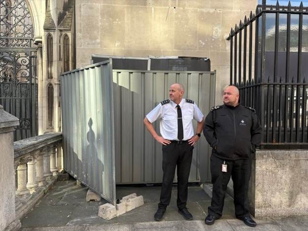 Two security guards, one in a white shirt and black tie with a badge, the other in a black jacket with a badge, standing near a metal storage container and black iron fence. The background includes a stone building with arched windows and decorative railings.