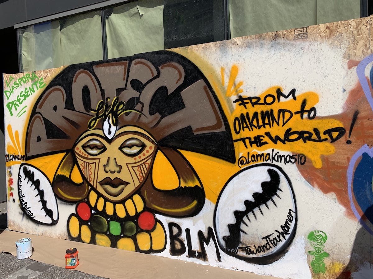 mural painted on a street wall representing a Black woman with a big gold, red, black, and green bead necklace. We can read “Protect Life” in her hair and “From Oakland to the world”, and “BLM” on the right of the painting.