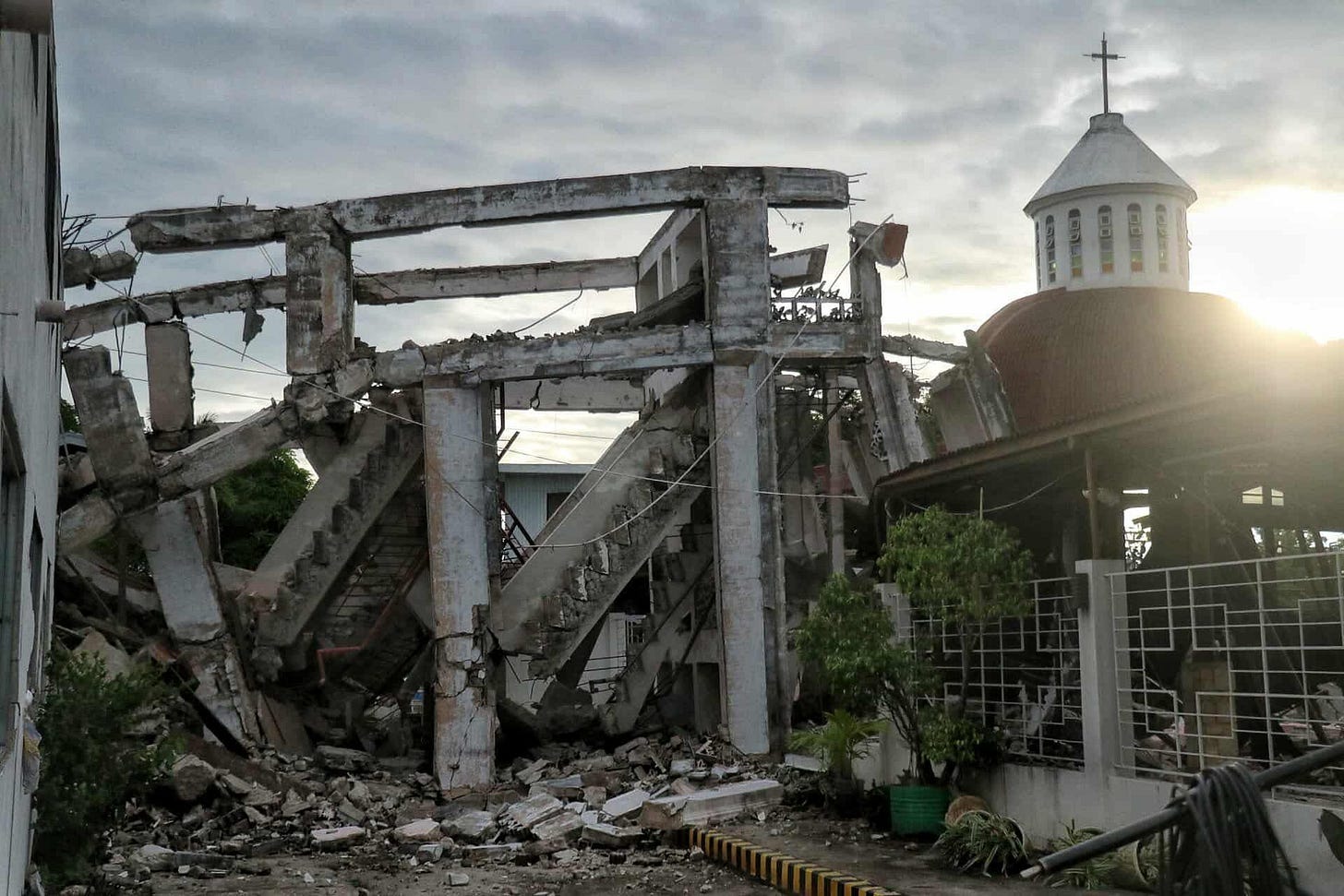 Damaged church and collapsed concrete building after an earthquake in the Philippines, with rubble scattered on the ground and the sun setting in the background. Damaged church and collapsed concrete building after an earthquake in the Philippines, with rubble scattered on the ground and the sun setting in the background.
