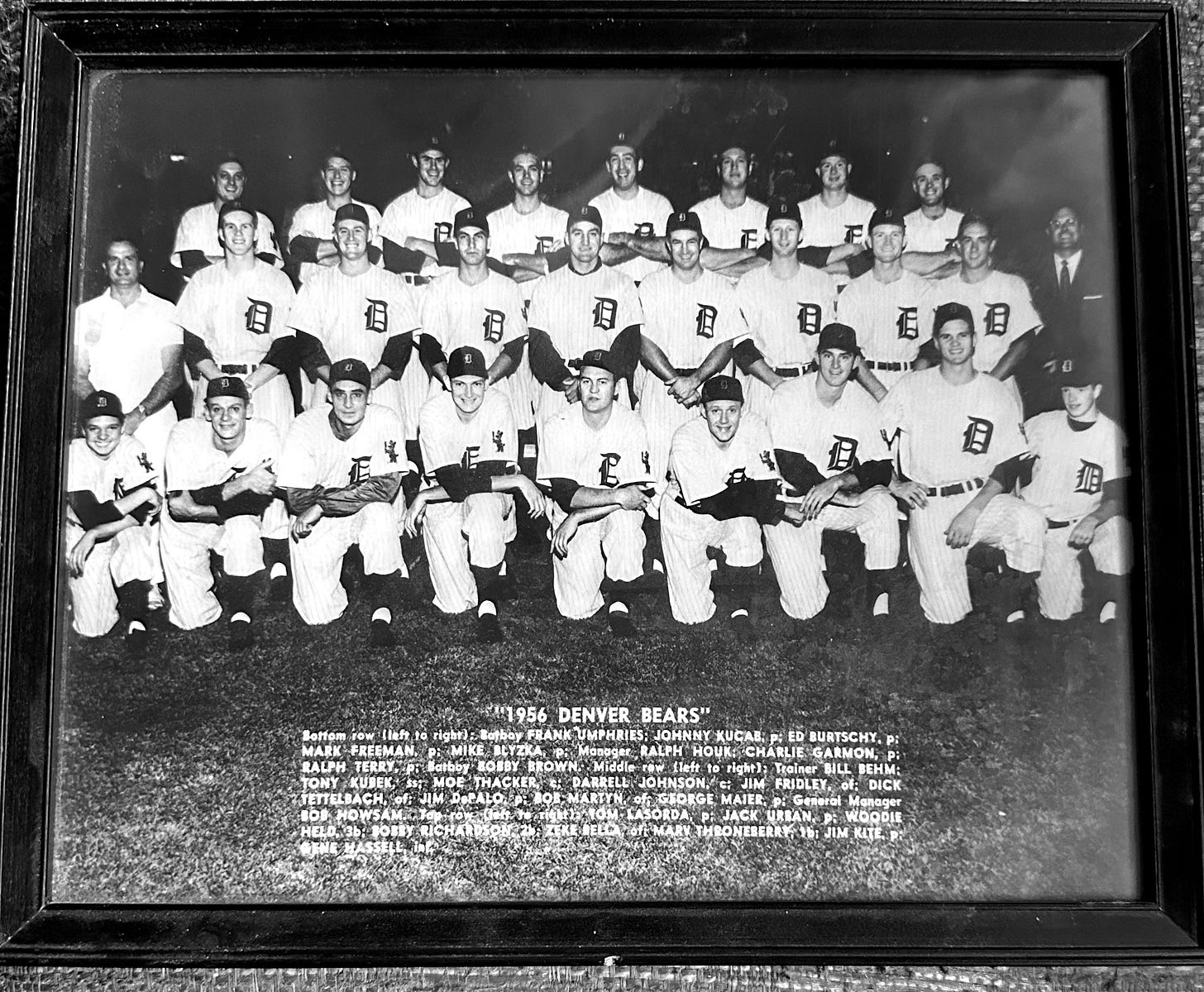 Monochrome 1956 Denver Bears team photo. Team members above, player names below. The photo caption reads: "“1956 DENVER BEARS" Bottom row (left to right): Batboy FRANK UMPHRIES; JOHNNY KUCAB, P; ED BURTSCHY, P; MARK FREEMAN, P; MIKE BLYZKA, P; Manager RALPH HOUK; CHARLIE GARMON, P; RALPH TERRY, P; Batboy BOBBY BROWN. Middle row (left to right); Trainer BILL BEHM; TONY KUBEK, S; MOE THACKER, C; DARRELL JOHNSON, C; JIM FRIDLEY, OF; DICK TETTELBACH, OF; JIM DePALO, P; BOB MARTYN, OF, GEORGE MAIER, P. General Manager BOB HOWSAM. Top row (left to right); TOM LASORDA, P; JACK URBAN, P; WOODIE HELD, 3B; BOBBY RICHARDSON, 2B; ZEKE BELLA, OF; MARV THRONEBERRY 1B; JIM KITE, P"