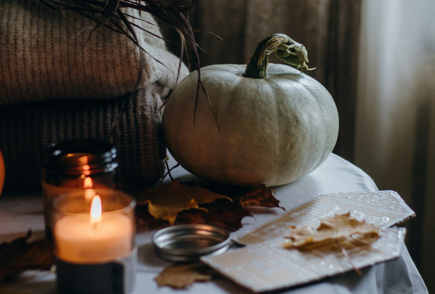Warm autumn still life featuring a white pumpkin, lit candle, scattered fall leaves, and soft brown knit blanket.