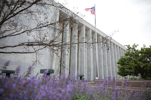 A flag on a pole waves in front of an off-white building with columns.
