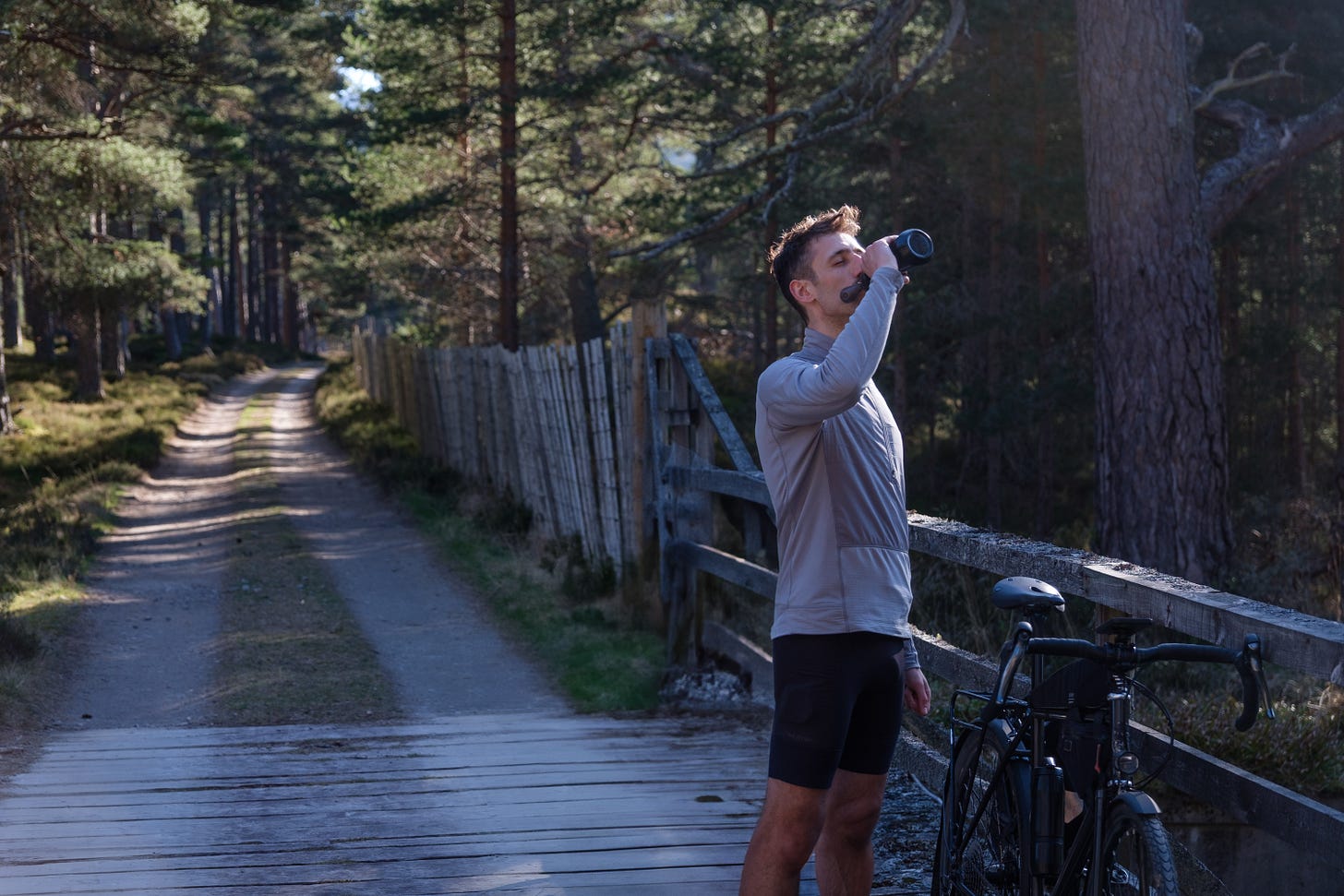 A person stands alone at the edge of a forest facing the sky with their eyes closed. They drink water from a bottle while a stream of light fills their face with a warm glow.