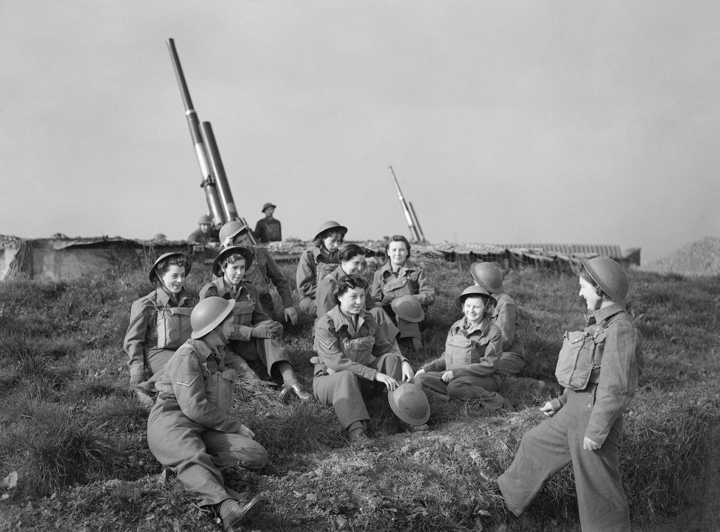 About a dozen women in British Army battledress and steel helmets sit and stand on grass beside an anti-aircraft gun emplacement, with two gun barrels raised behind them. About a dozen women in British Army battledress and steel helmets sit and stand on grass beside an anti-aircraft gun emplacement, with two gun barrels raised behind them.