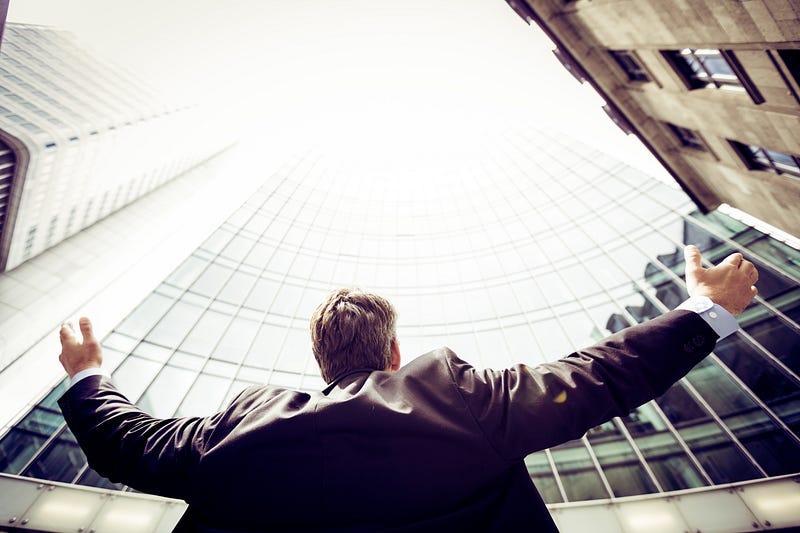 Business person standing outside building with arms out, looking up