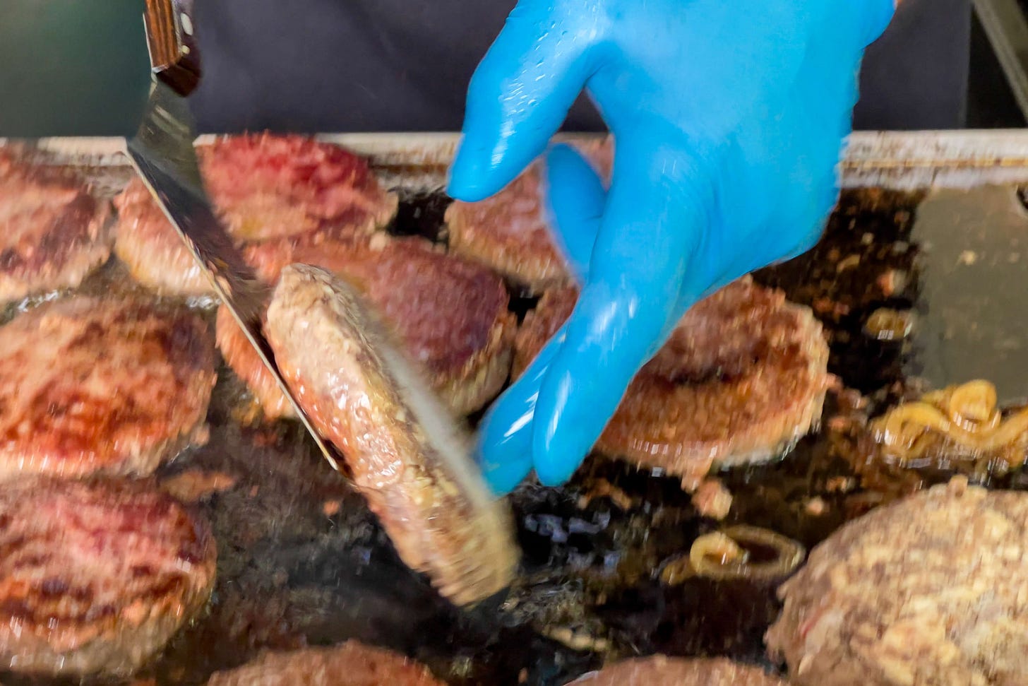 Nahaufnahme einer Grillplatte voller brutzelnder Hamburger-Patties aus dem Fleisch japanischer Wagyu-Rinder. Eine Hand mit blauer Einweghandschuhen hebt mit einer Metallkelle einen halb durchgebratenen Patty an. Fett und Fleischsaft schäumen auf der heissen, dunklen Grillfläche, daneben liegen angeröstete Zwiebeln.