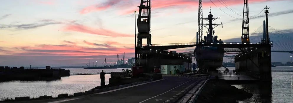 A industrial port or shipyard scene photographed at dusk or dawn, featuring large cranes and a vessel docked alongside a pier. The dramatic sky displays vivid pink and purple hues from the setting or rising sun, reflecting off the calm water. A curved road runs along the waterfront in the foreground, with port infrastructure silhouetted against the colorful sky. A industrial port or shipyard scene photographed at dusk or dawn, featuring large cranes and a vessel docked alongside a pier. The dramatic sky displays vivid pink and purple hues from the setting or rising sun, reflecting off the calm water. A curved road runs along the waterfront in the foreground, with port infrastructure silhouetted against the colorful sky.