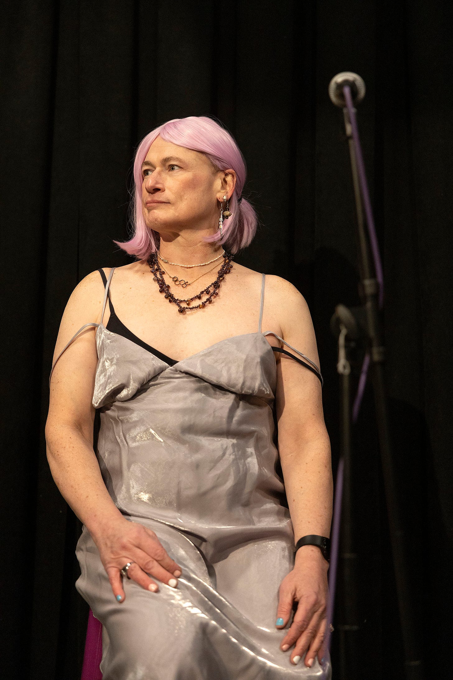 Photo of Davi Gray, seated alone on a stage. She is wearing a silver gown, several necklaces, pink hair, and nails painted in the colors of the trans pride flag.