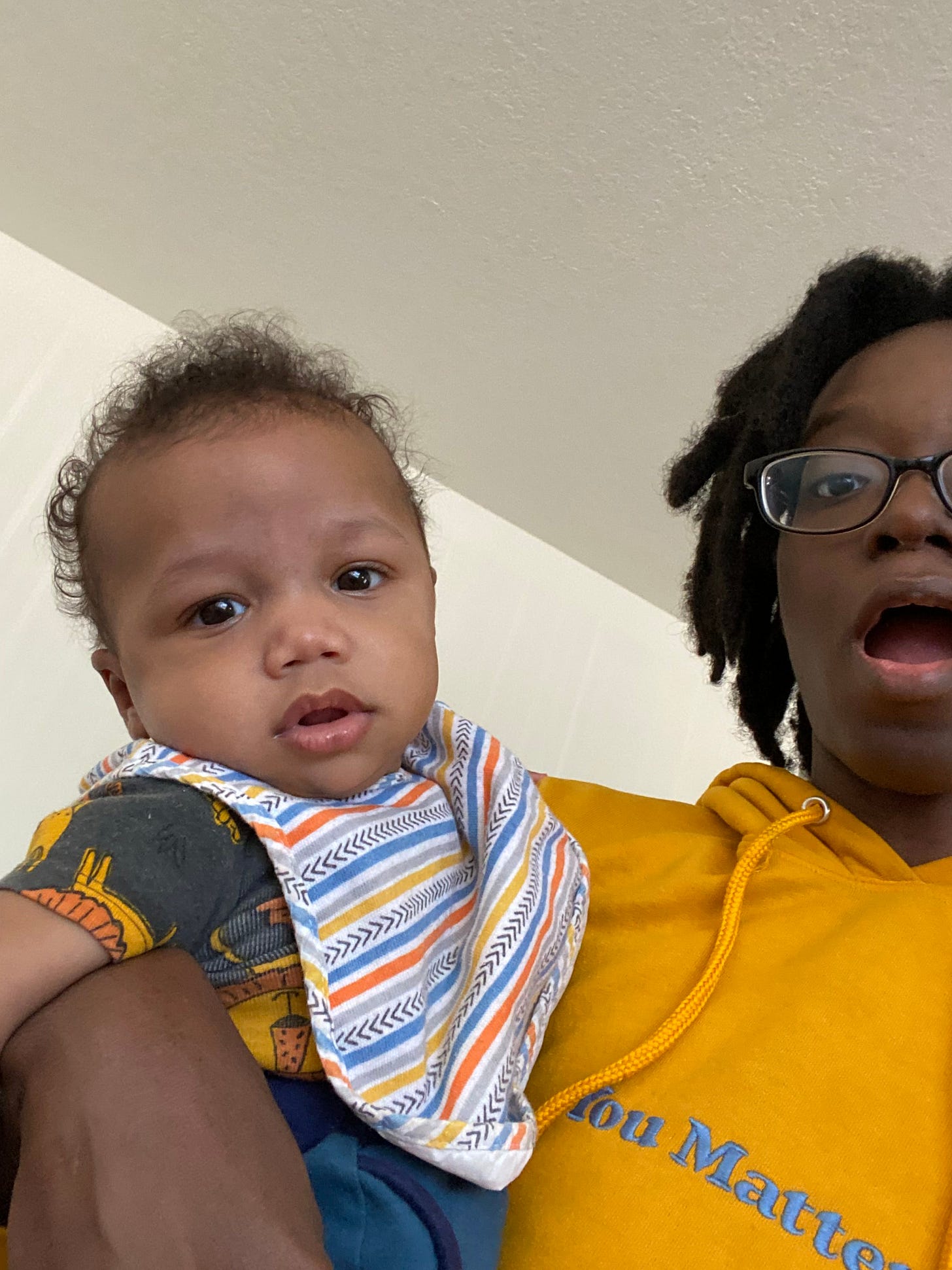 a Black person with freeform locs wearing black glasses, a yellow hoodie that says “You Matter” in blue stitching, holding a light-skinned baby.
