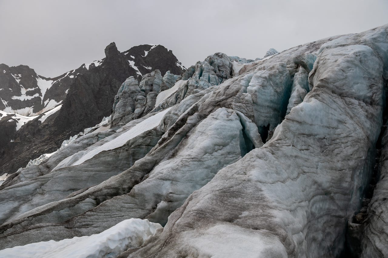 Standing low on the buckled ice of an icefall, looking across ridges of dirty grey-blue glacier ice split by crevasses, one dark slot opening just right of center. The ice rises in pressure ridges toward pale blue seracs above, and beyond them a black rock peak with snow-filled gullies stands against a flat grey sky. Bands of rock dust streak and darken the ice everywhere except a few clean white patches of snow.