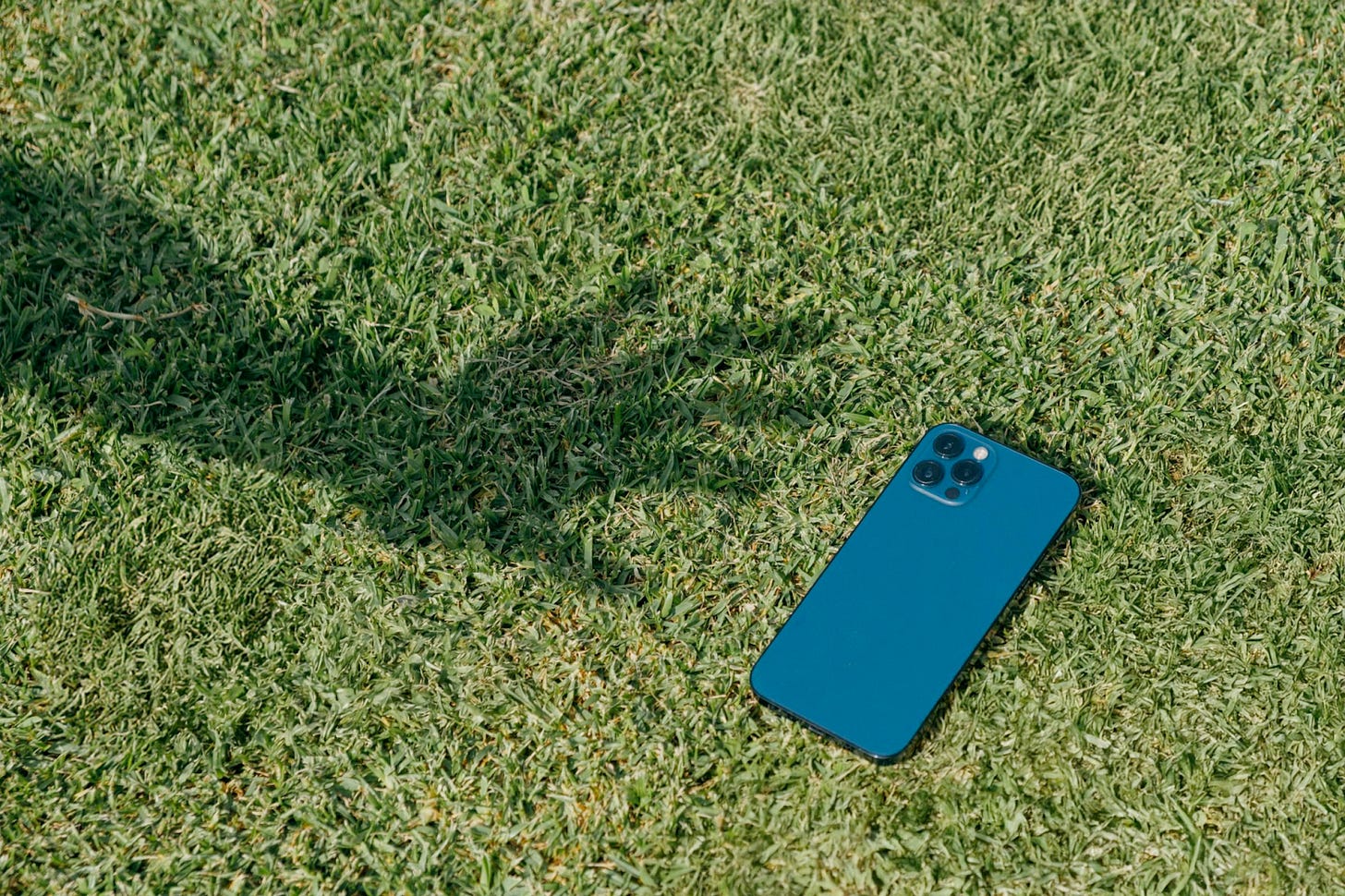 A teal smartphone with triple rear cameras lies on green grass, next to the shadow of an outstretched arm.