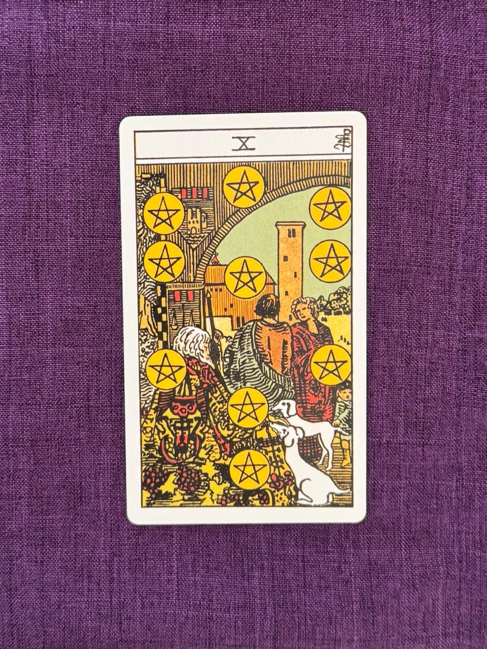 tarot card on a purple background: the ten of pentacles An old, white-haired man wearing an ornately embroidered robe, sits with his two white dogs at his feet. A younger couple stands nearby with a small child. The man and his family gather in the courtyard of a large castle. On the archway are family emblems and flags. Around them, there are ten yellow circles, each with a five pointed star in them. tarot card on a purple background: the ten of pentacles An old, white-haired man wearing an ornately embroidered robe, sits with his two white dogs at his feet. A younger couple stands nearby with a small child. The man and his family gather in the courtyard of a large castle. On the archway are family emblems and flags. Around them, there are ten yellow circles, each with a five pointed star in them.