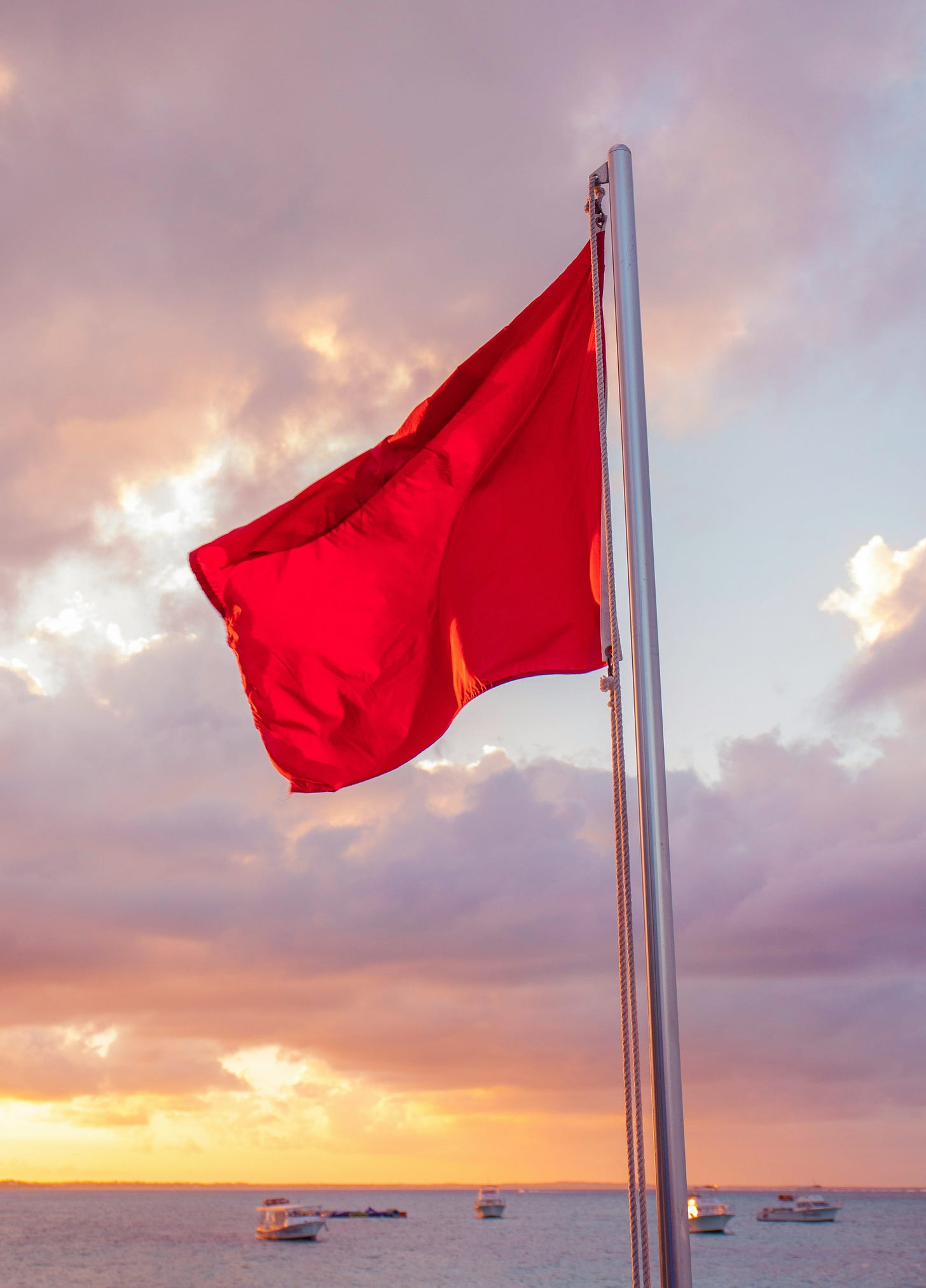 A bright red flag flutters on a pole against a dramatic sky at sunset. The background shows soft purple and orange clouds over a calm ocean with several boats anchored in the distance. A bright red flag flutters on a pole against a dramatic sky at sunset. The background shows soft purple and orange clouds over a calm ocean with several boats anchored in the distance.