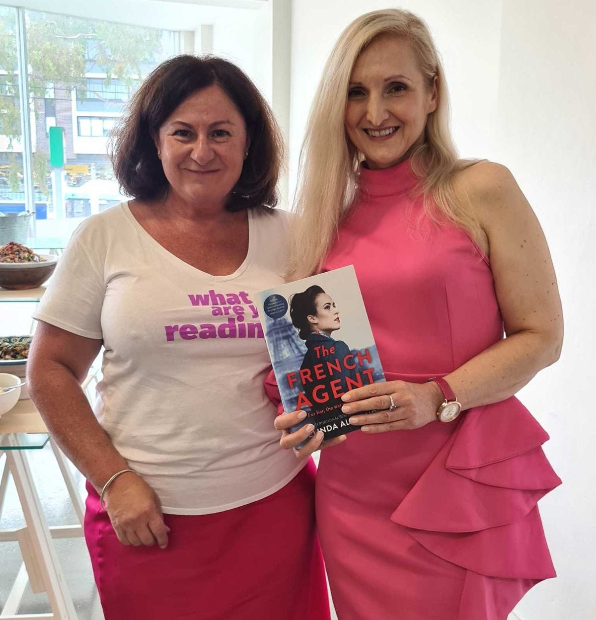 Two women standing next to each other smiling. The woman on the left has short brown hair and where's a white t-shirt that says 'What are you Reading?' And a pink skirt. The woman on the right with long blonde hair is Belinda Alexandra in a pink halter dress and holding a copy of her book The French Agent.