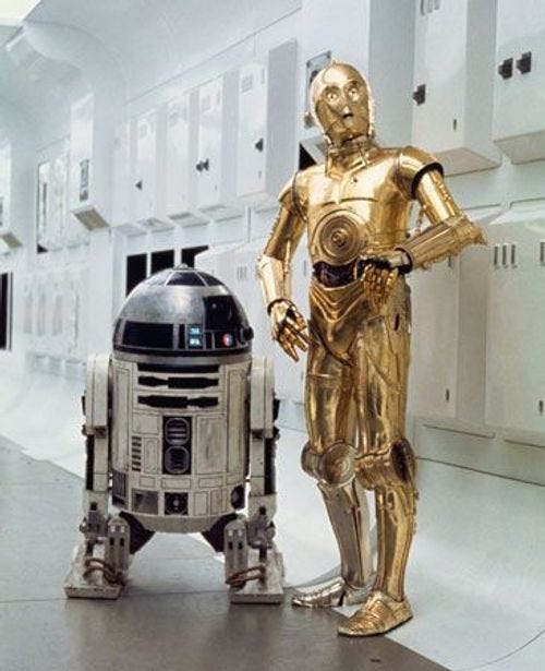The Star Wars droids R2-D2 and C-3PO stand by each other in a white hallway. The Star Wars droids R2-D2 and C-3PO stand by each other in a white hallway.