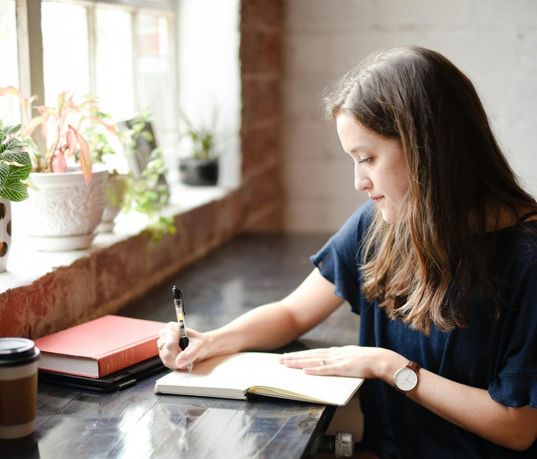 woman sitting in front of black table writing on white book near window woman sitting in front of black table writing on white book near window