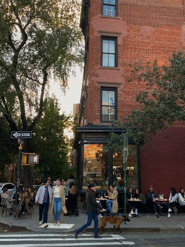 This may contain: people are walking across the street in front of a brick building with an awning This may contain: people are walking across the street in front of a brick building with an awning