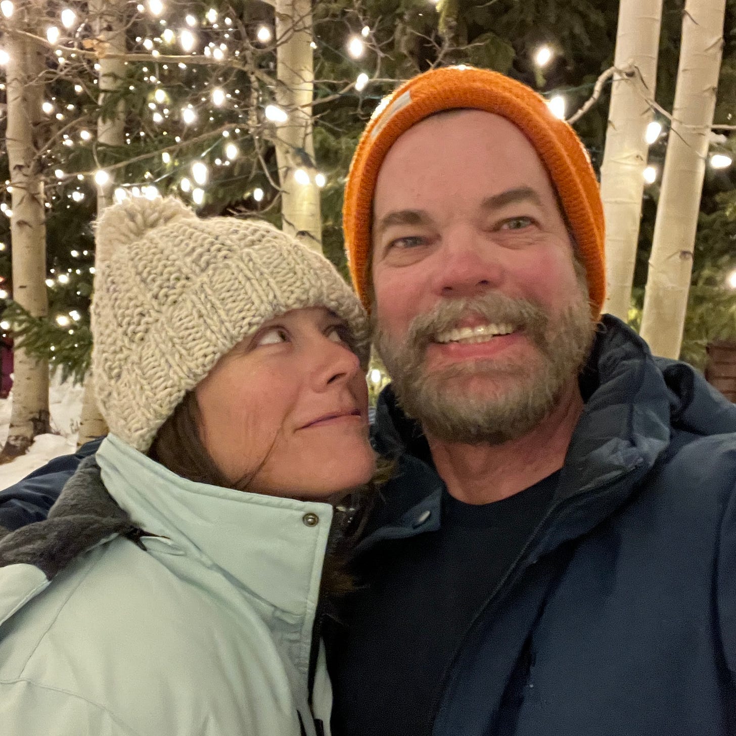 Brett in a cream knit winter hat and her husband wearing an orange hat outside surrounded by twinkle lights