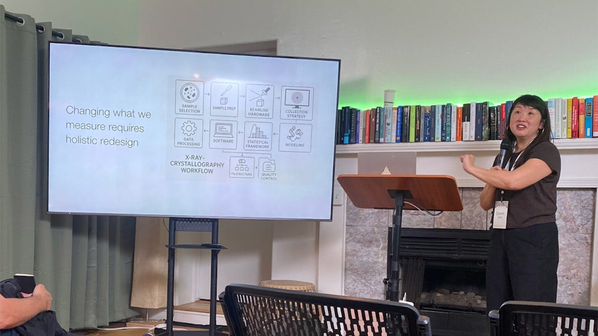 A woman stands at a podium presenting a slide about holistic redesign and X-ray crystallography workflow to an audience in a room with bookshelves and a fireplace.