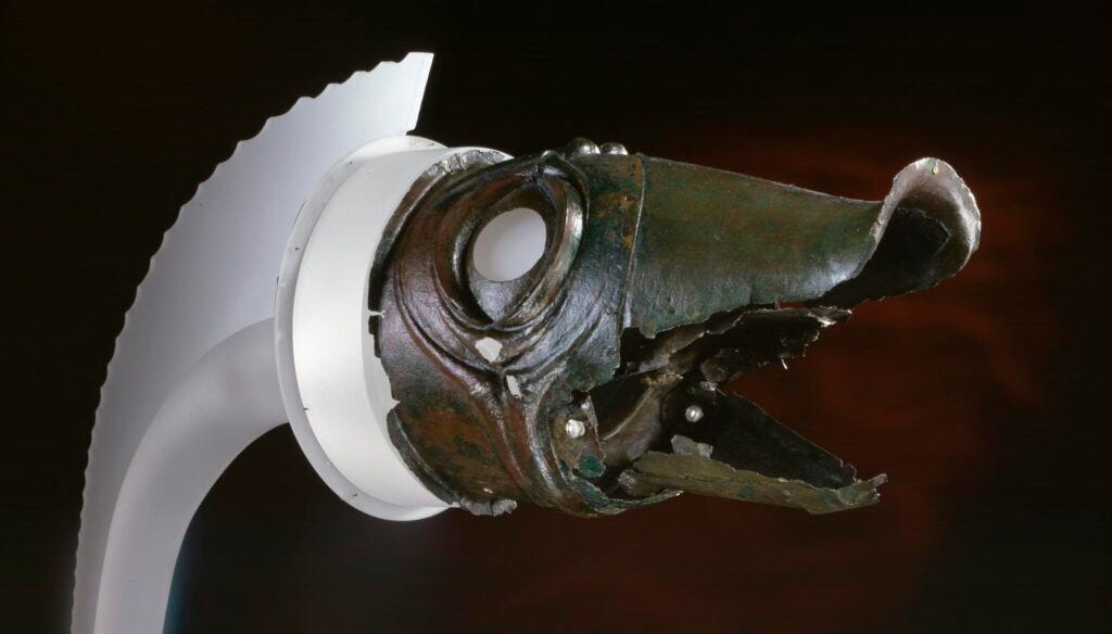 A close-up profile view of the ancient bronze and brass Deskford Carnyx head, a Celtic war-horn crafted in the detailed shape of a stylized wild boar with an open, hollow mouth, prominent circular eyes, and a ribbed crest along its spine against a dark background.