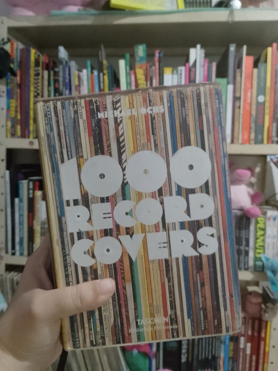 A imagem destaca uma pessoa segurando em primeiro plano o livro "1000 Record Covers", de autoria de Michael Ochs e publicado pela editora Taschen. A capa do livro apresenta uma estampa vertical colorida que simula a visão de dezenas de lombadas de discos de vinil enfileirados em uma coleção. Centralizado na capa, o título do livro aparece em letras brancas, grandes e estilizadas. Ao fundo, uma estante de madeira clara serve de cenário, exibindo diversas prateleiras repletas de livros e quadrinhos coloridos organizados na vertical, o que complementa o ambiente de leitura e coleção.