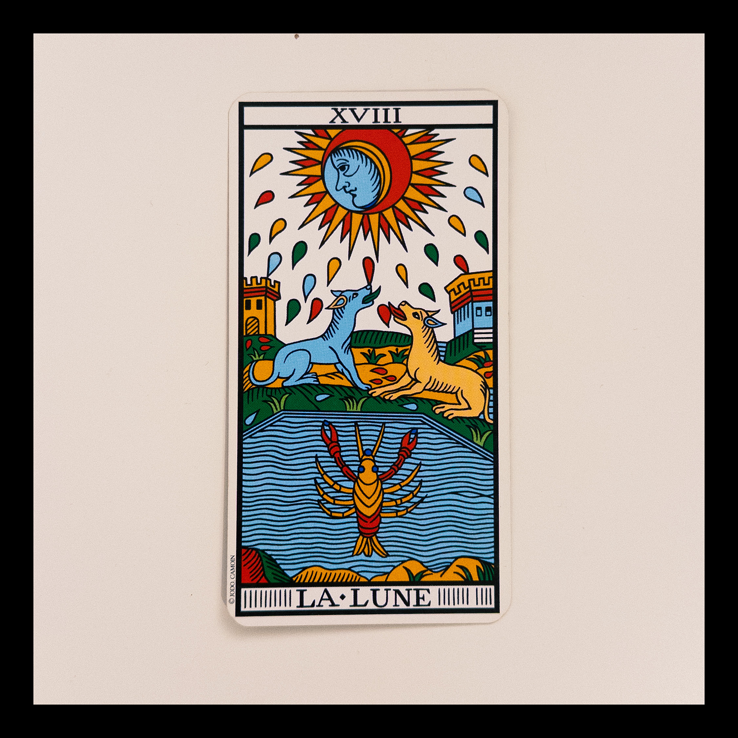 Carta del tarot “La Luna (XVIII)” del Tarot de Marsella, versión restaurada por Philippe Camoin y Alejandro Jodorowsky. Muestra una luna con rostro azul que observa hacia un lado, rodeada de gotas de colores. Abajo, un cangrejo emerge de un estanque. Dos torres y dos cánidos —uno rojo, uno azul— completan la escena. La imagen sugiere intuición, confusión emocional, ciclos inconscientes y el paso por una etapa incierta pero fértil.