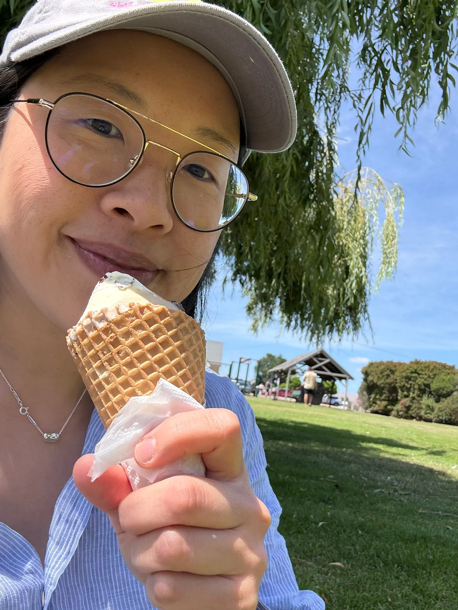 Sonya smiling while eating an ice cream cone in a park, wearing glasses, a grey cap and a blue and white striped shirt.