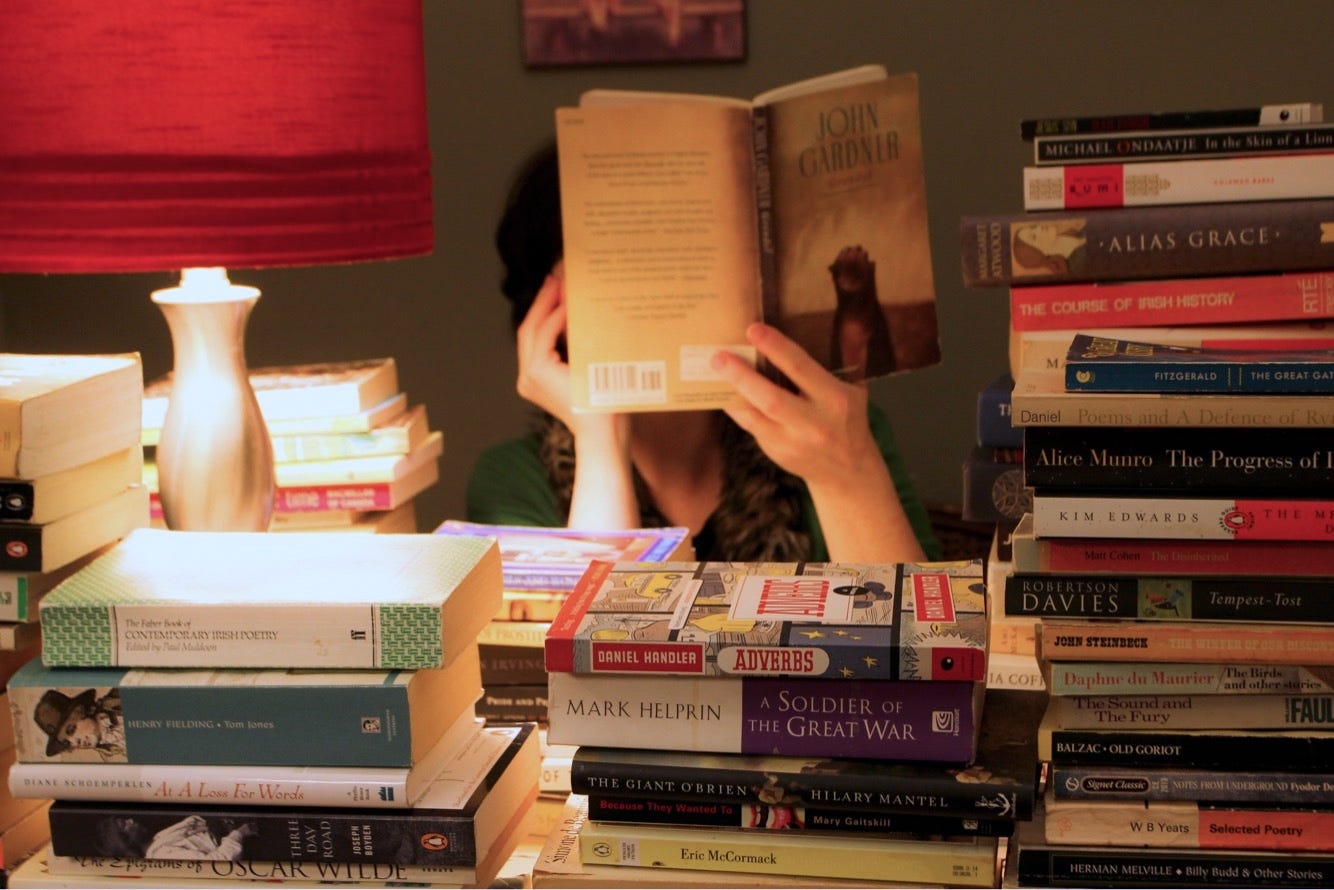 Behind various stack of books at different heights, a red lamp illuminates the scene. I am sitting behind the desk with about 100 books on it, reading John Gardnener's novel, "Grendel" as a nod to Beowulf, one of the oldest tales in English storytelling. This was a selfie for a Flickr project. Photo credit: me. Behind various stack of books at different heights, a red lamp illuminates the scene. I am sitting behind the desk with about 100 books on it, reading John Gardnener's novel, "Grendel" as a nod to Beowulf, one of the oldest tales in English storytelling. This was a selfie for a Flickr project. Photo credit: me.